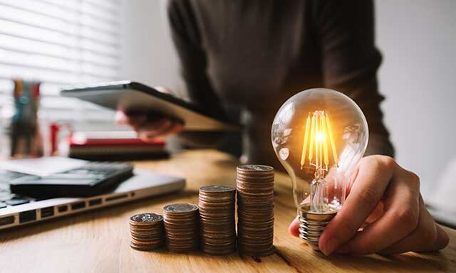 Frau hält eine Glühlampe, daneben Münzstapel